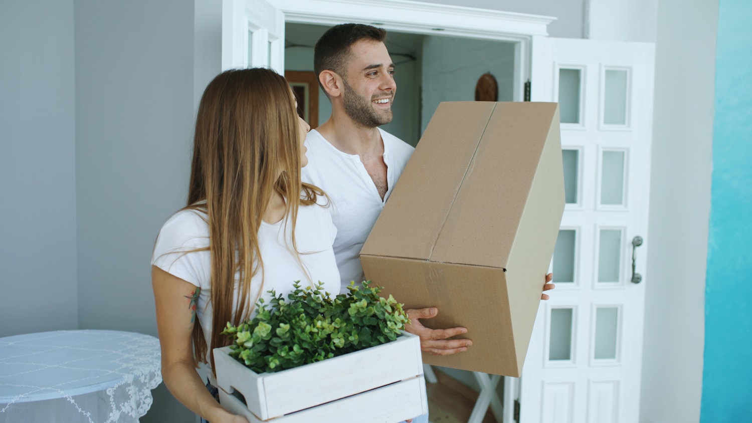 A happy couple carrying a moving box and plant into their new home